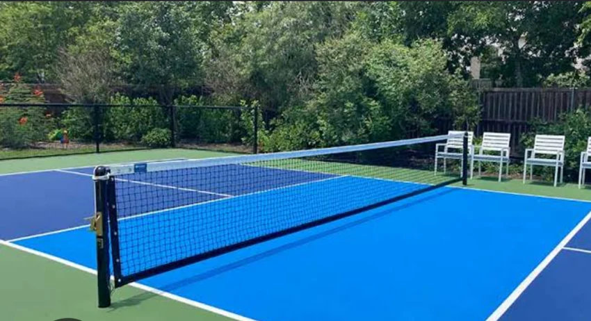 Blue pickleball court with a green surround, net, fencing, and courtside chairs